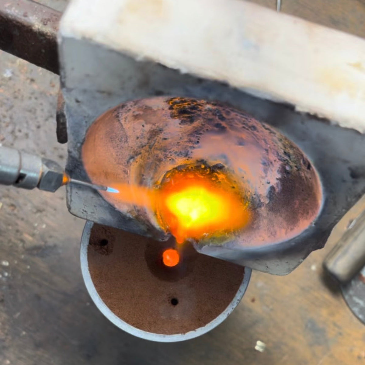 A close-up of a metal object being heated in a forge or torch, with bright orange and yellow molten light visible against a dark industrial background.