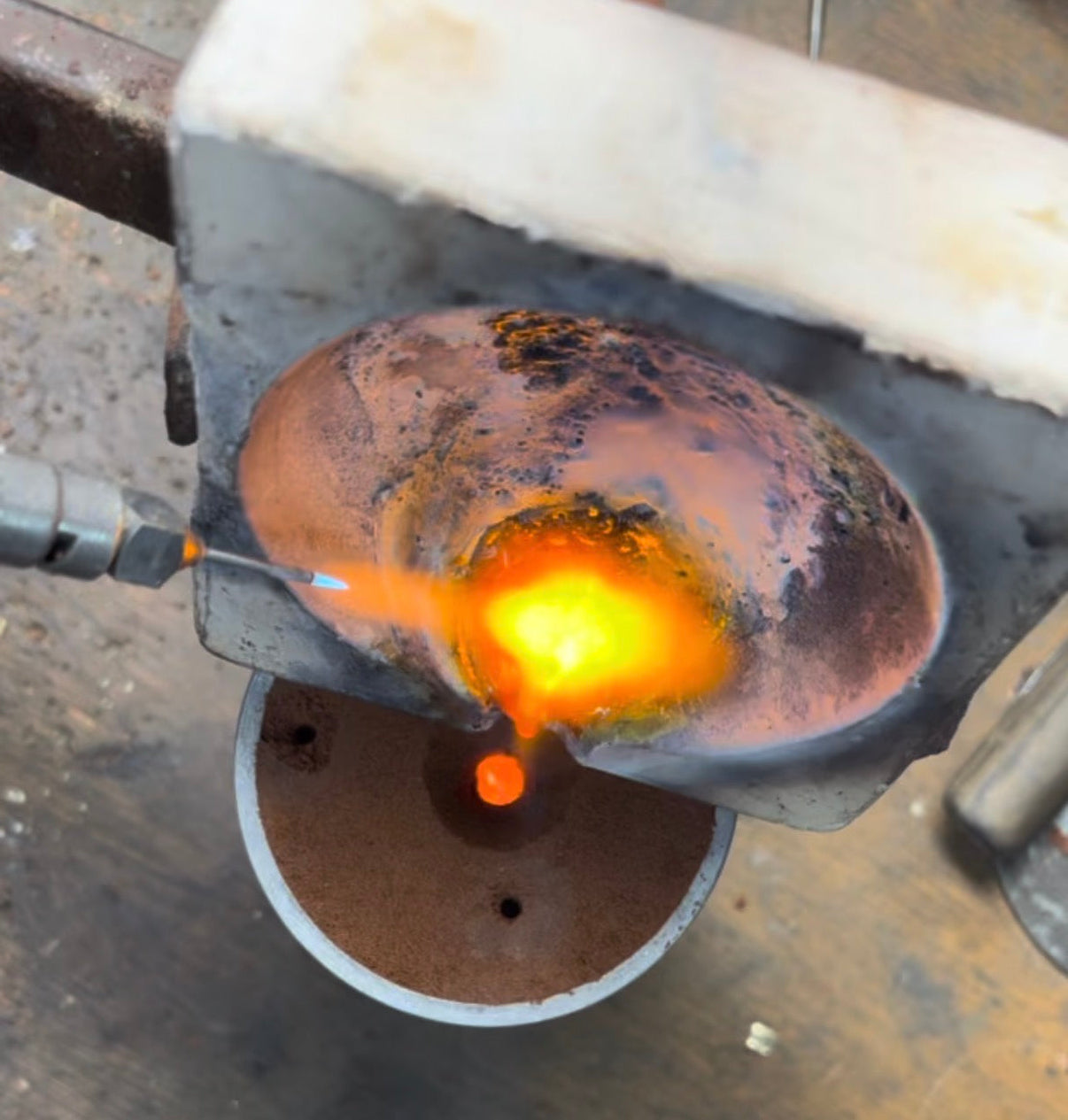 A close-up of a metal object being heated in a forge or torch, with bright orange and yellow molten light visible against a dark industrial background.
