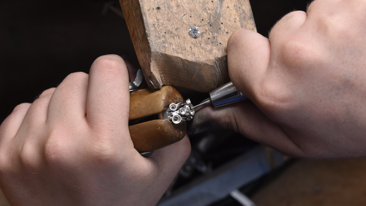Close-up of a jeweler's hands using a diamond-setting tool on a small metal ring cluster held in a wooden clamp, with a wooden mallet in the background.