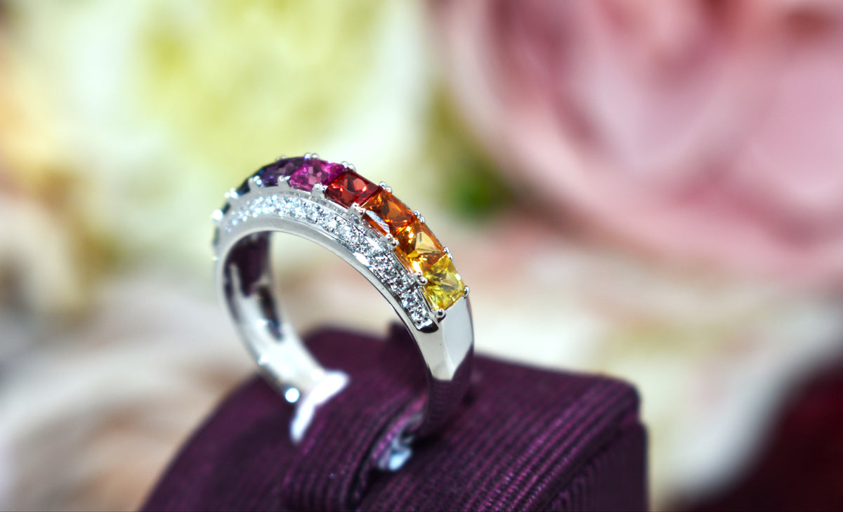 A silver band ring featuring a row of colorful gemstones in pink, orange, yellow, and green, displayed on a dark purple velvet ring box with a blurred floral background.