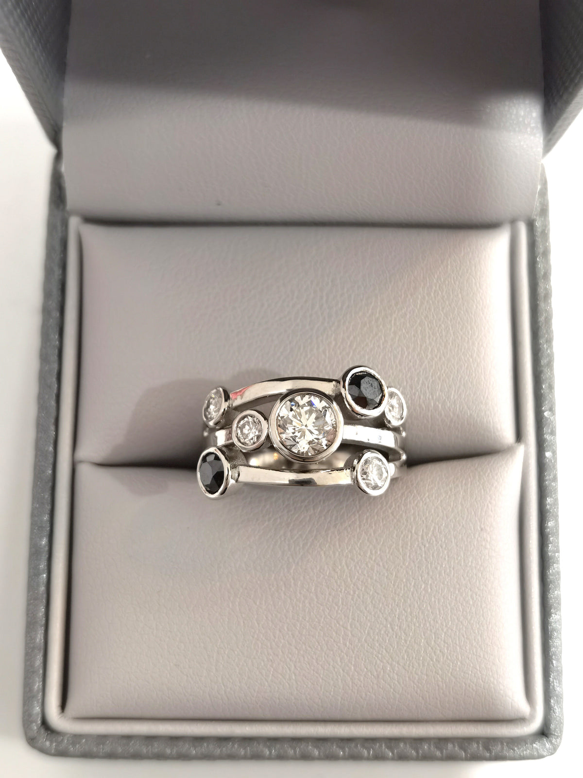 A silver-toned multi-band ring with a central round clear gemstone and smaller accent stones, displayed on a gray ring box cushion.
