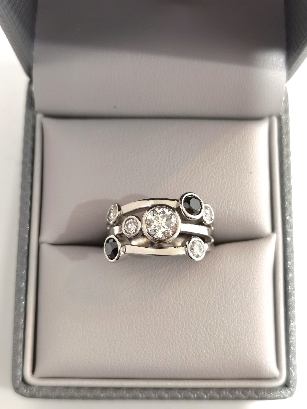 A silver-toned multi-band ring with a central round clear gemstone and smaller accent stones, displayed on a gray ring box cushion.