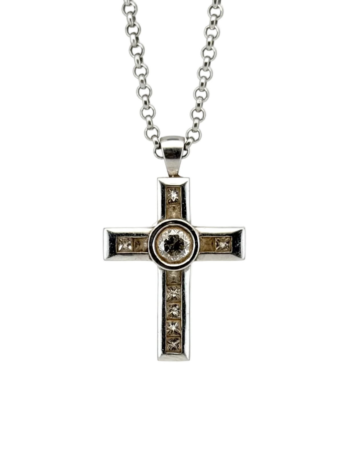 A silver cross pendant necklace with a central round dark gemstone and small clear stones along the arms, suspended from a silver chain on a white background.